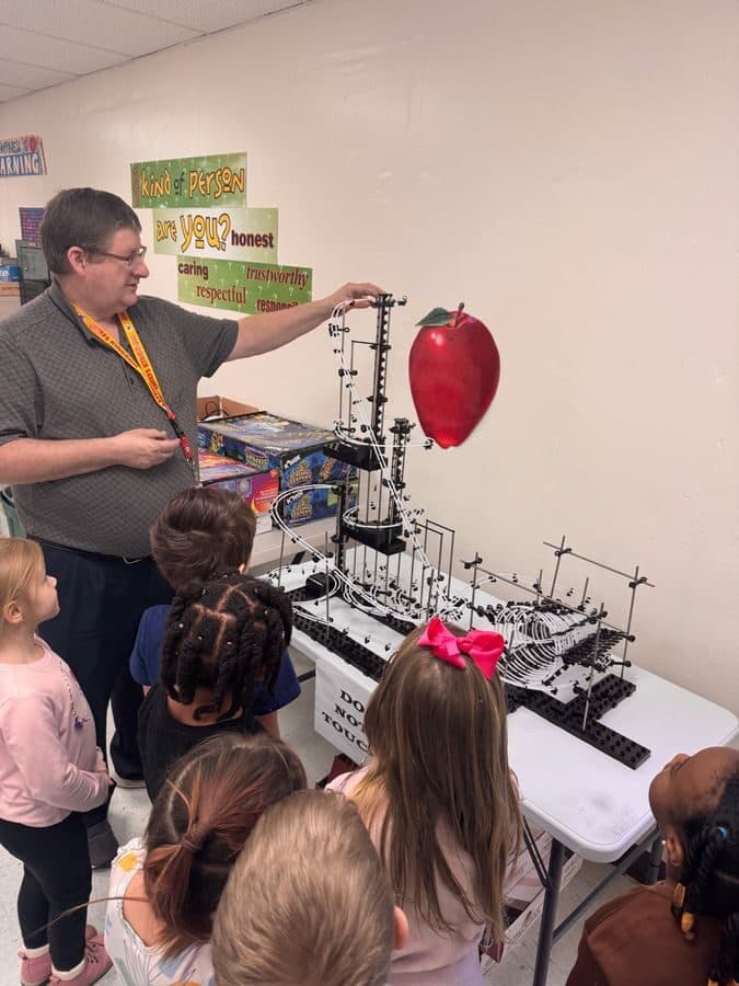 An elementary teacher demonstrates a hands-on science project to students gathered around a table