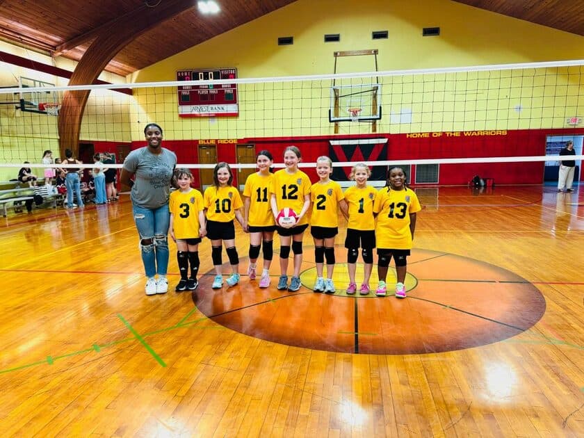An elementary volleyball team poses together on the court in the Warriors gym